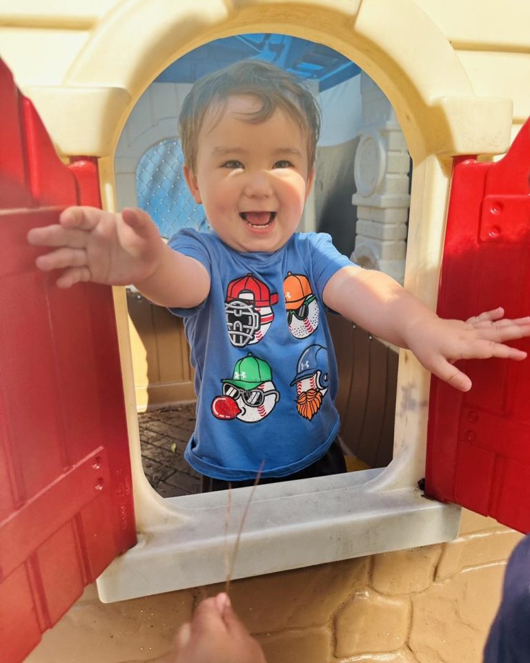 Smiling boy opening playhouse window during outdoor play at Ardent Preschool & Daycare – Trace Crossings